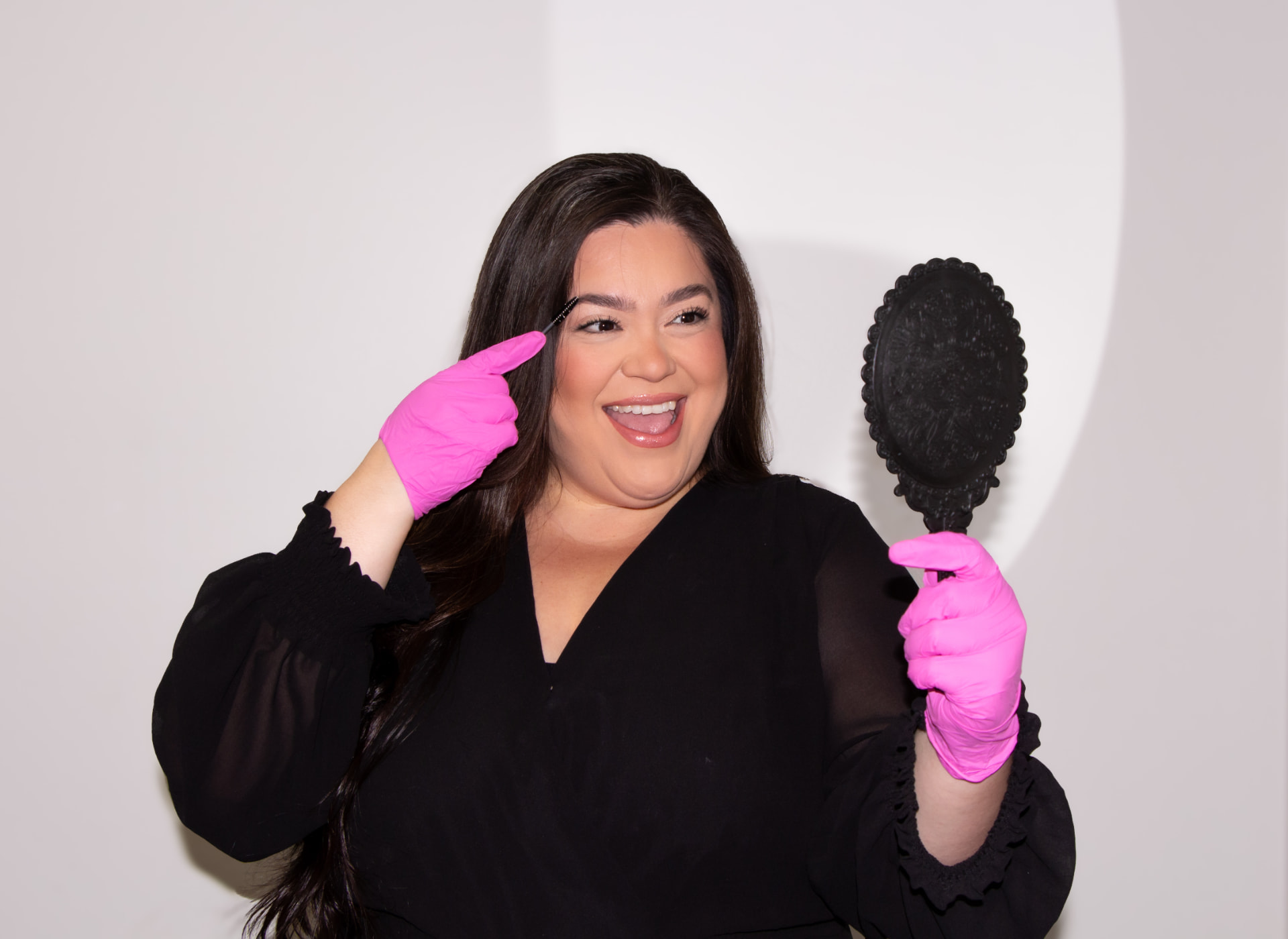 Jaquie smiling in pink gloves pointing to her brows while holding a vintage handheld mirror